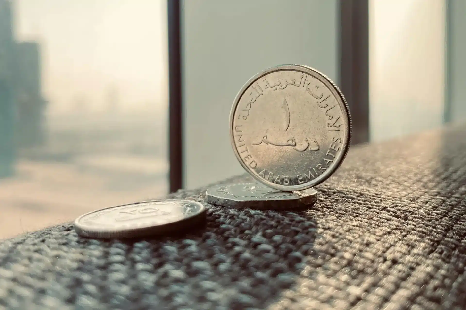 Close-up of UAE dirham coins on a window ledge with a blurred Dubai skyline behind