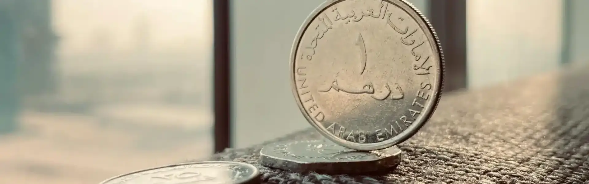 Close-up of UAE dirham coins on a window ledge with a blurred Dubai skyline behind